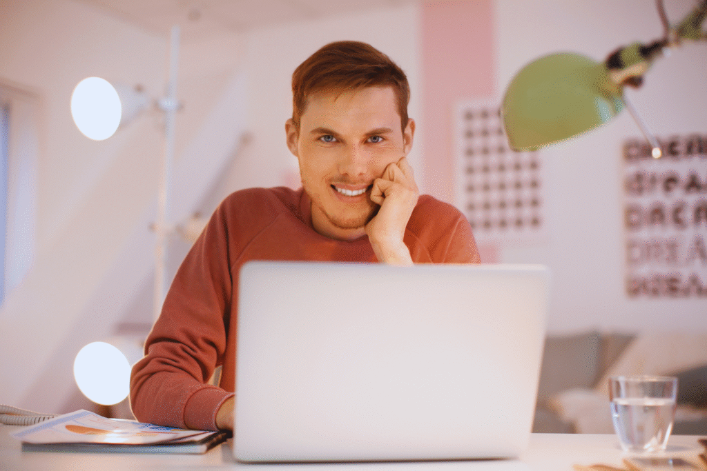 Um jovem de moletom rosa está sentado em sua mesa, sorrindo para a câmera com a mão no rosto e um laptop aberto à sua frente - um instantâneo ideal de o que é teletrabalho. Um copo de água, papéis e uma lâmpada verde completam a cena.