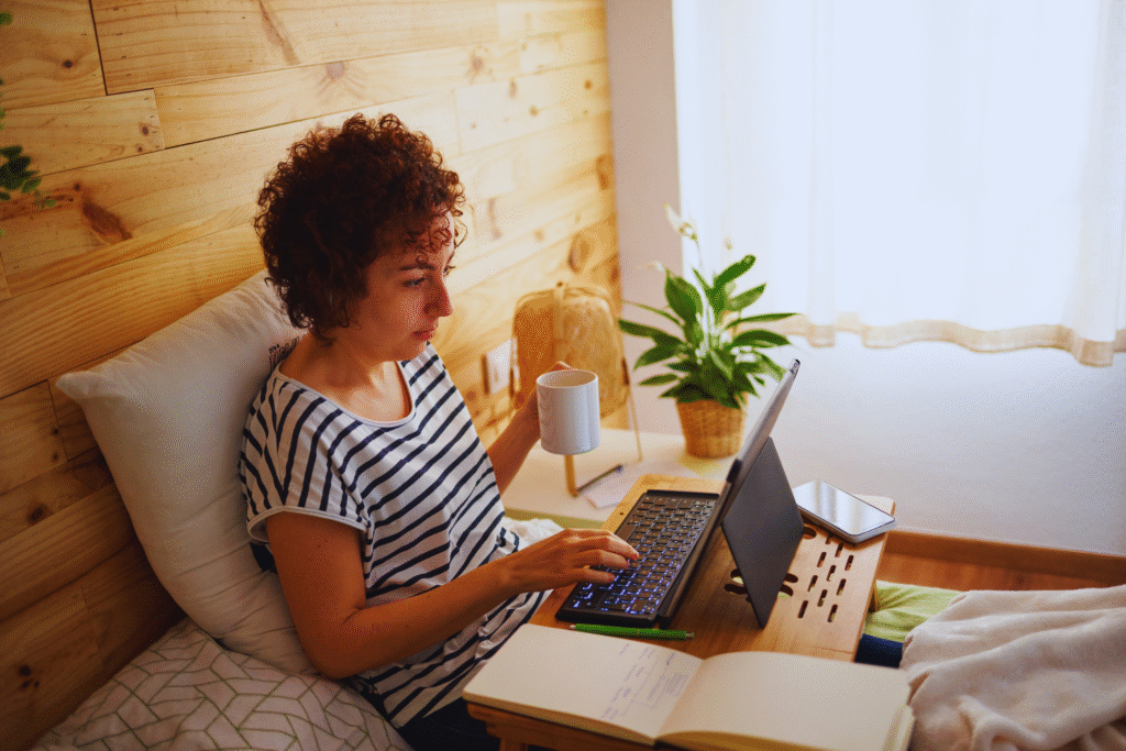 A woman with curly hair sits on a bed using a laptop on a wooden tray, embodying o que é teletrabalho. She wears a striped shirt, with a notebook and coffee mug nearby in her cozy, sunlit room with wooden walls and a potted plant.
