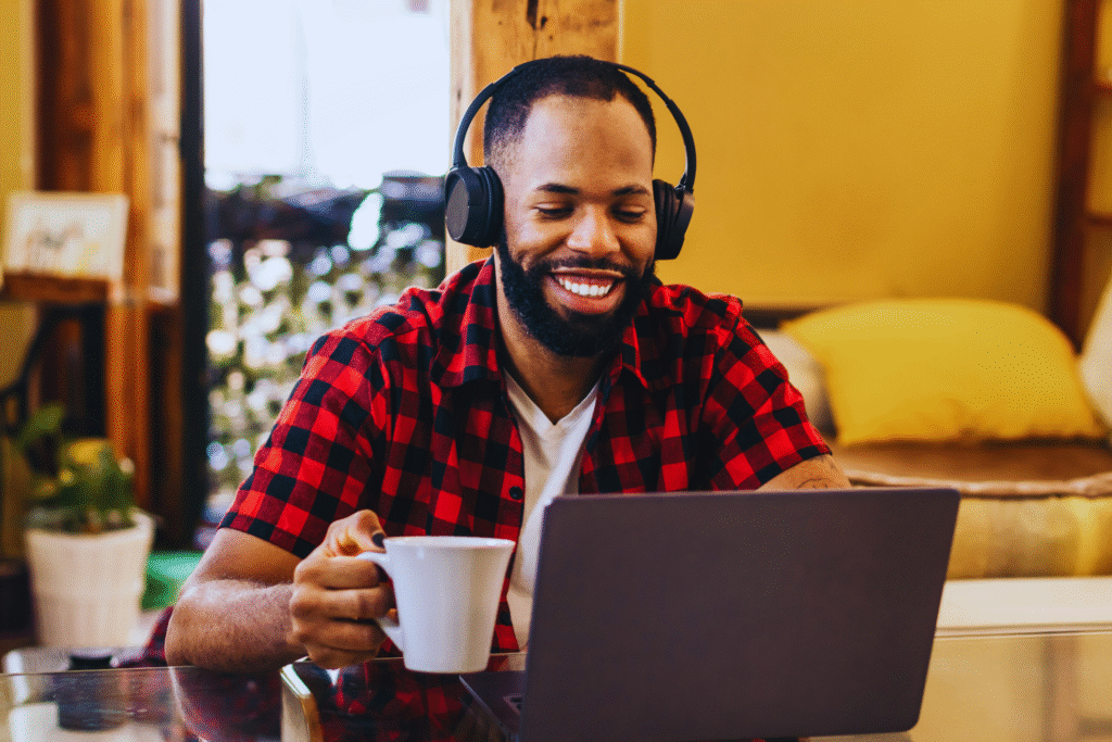 A man wearing headphones and a red plaid shirt sits at a table, smiling at his laptop screen while holding a white mug—enjoying the comfort of teletrabalho CLT in a cozy, well-lit living space with yellow cushions.
