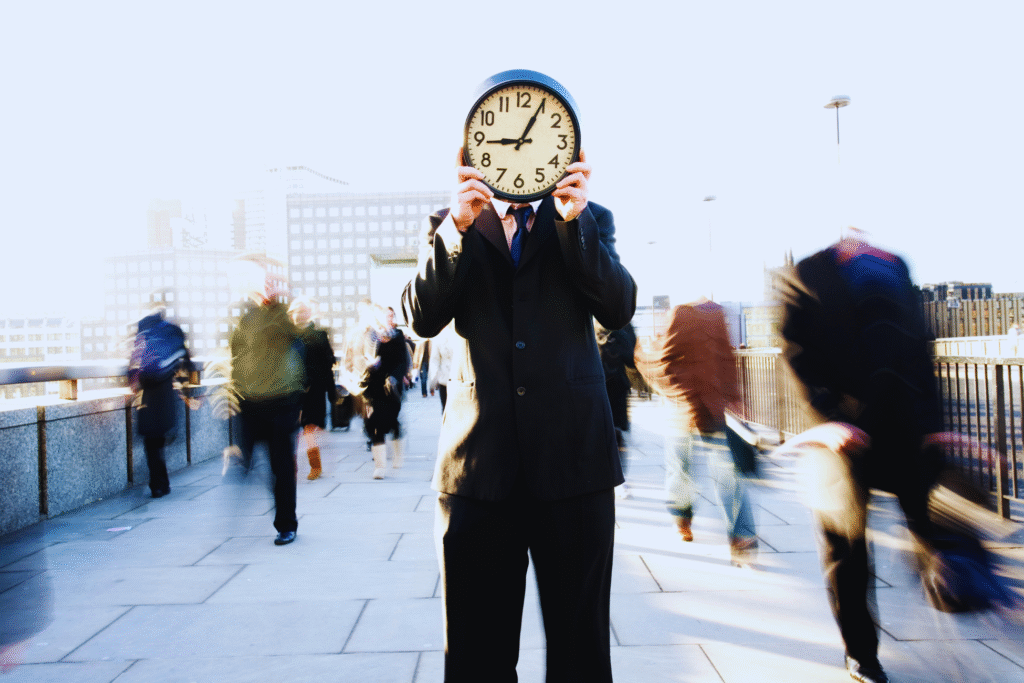 A person in a suit stands on a busy city sidewalk holding a clock in front of their face, while blurred people rush by—evoking the urgency of managing time, much like understanding o que é intervalo intrajornada.