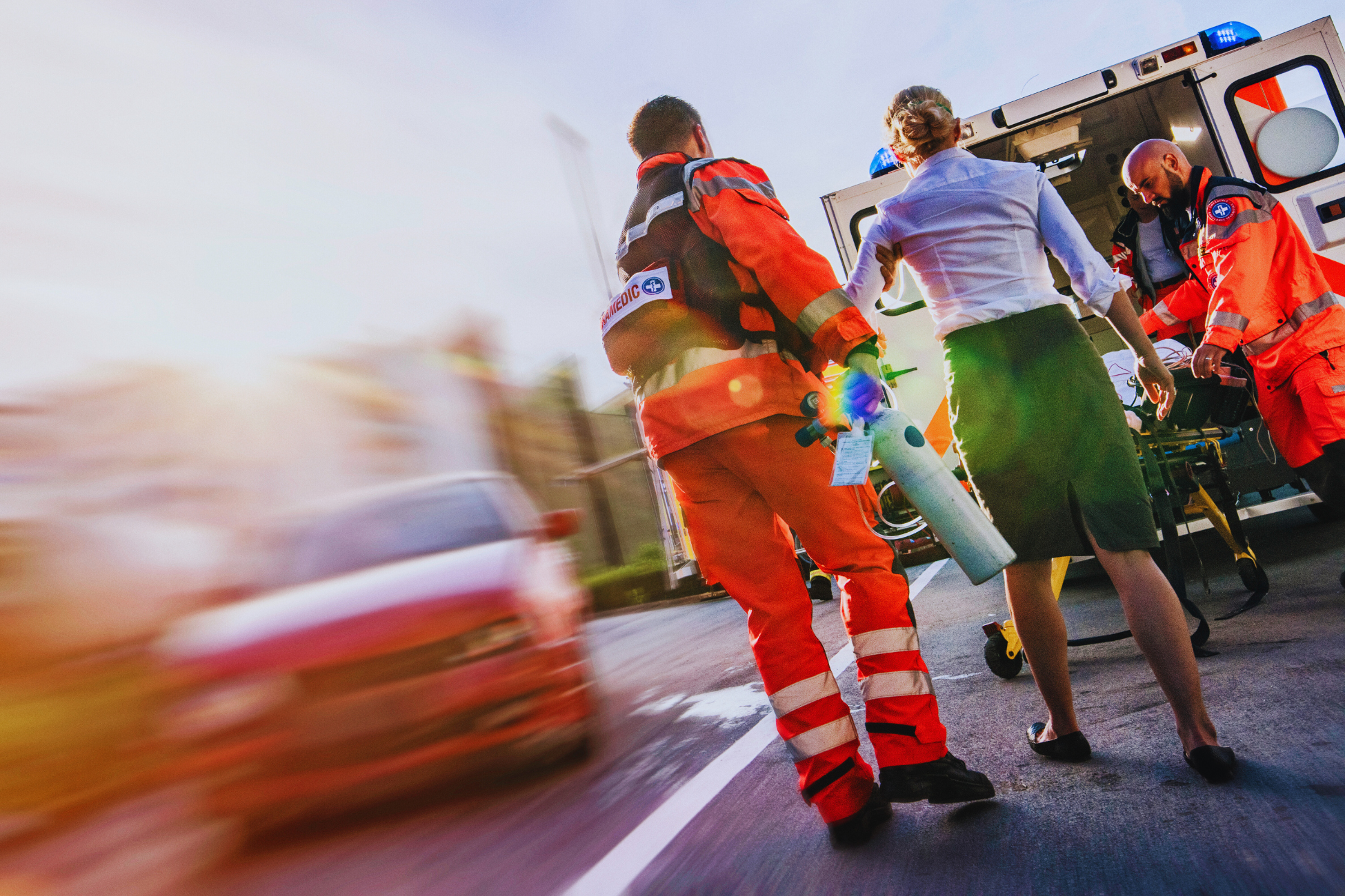 Emergency responders in orange uniforms assist a woman toward an ambulance after um acidente em serviço externo CAT, with a blurred red car and city backdrop, highlighting urgency and swift action.